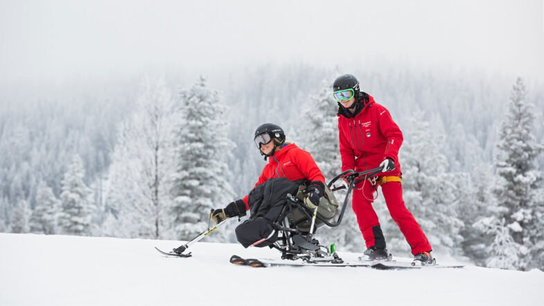 En person åker sitski med stöd av en skidlärare, båda ler. Snötäckta träd i bakgrunden