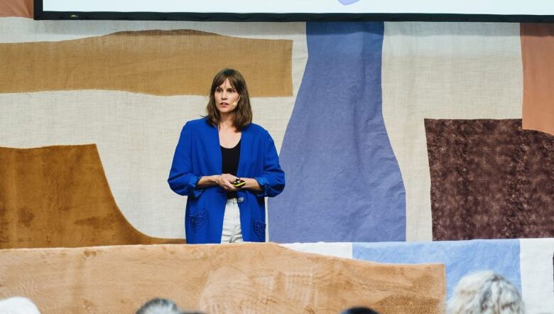 En kvinna med mörkbrunt hår iklädd vita byxor, svart linne och en blå kavaj står bakom ett bord och föreläser