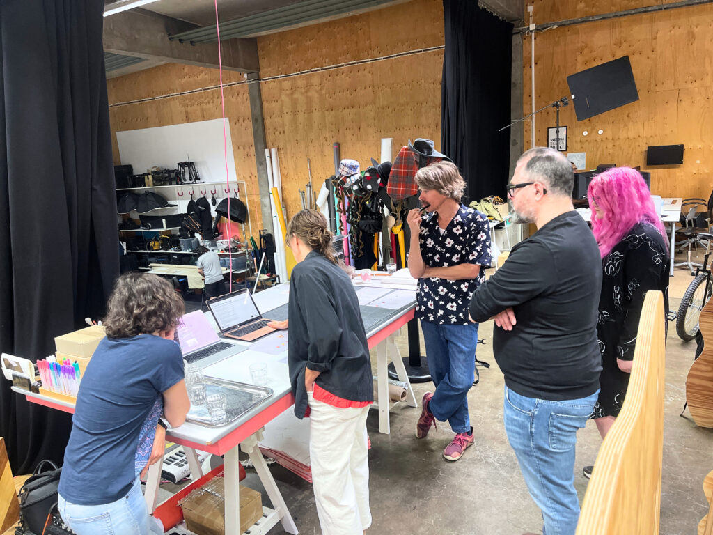 Fem personer står samlade runt ett bord i en kreativ studio. Två av dem arbetar vid laptops, medan de andra följer diskussionen. På bordet ligger papper, pennor och glas, och i bakgrunden syns rekvisita och verktyg