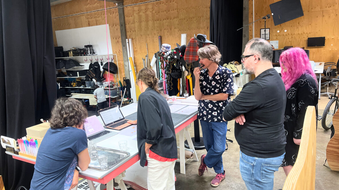 Fem personer står samlade runt ett bord i en kreativ studio. Två av dem arbetar vid laptops, medan de andra följer diskussionen. På bordet ligger papper, pennor och glas, och i bakgrunden syns rekvisita och verktyg