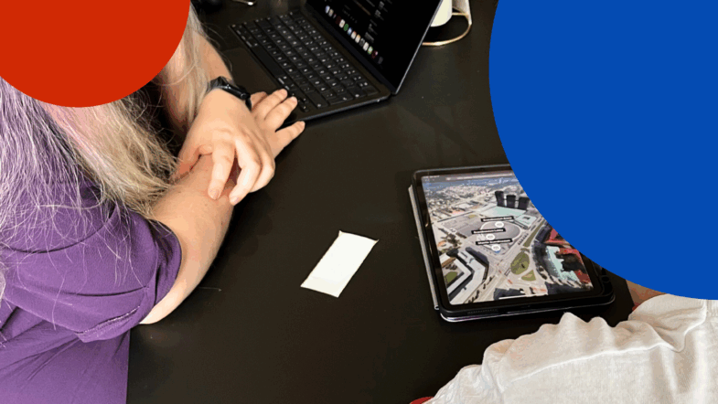 Två personer sitter vid ett bord och samarbetar. En bärbar dator visar text medan en surfplatta visar en stadskarta.