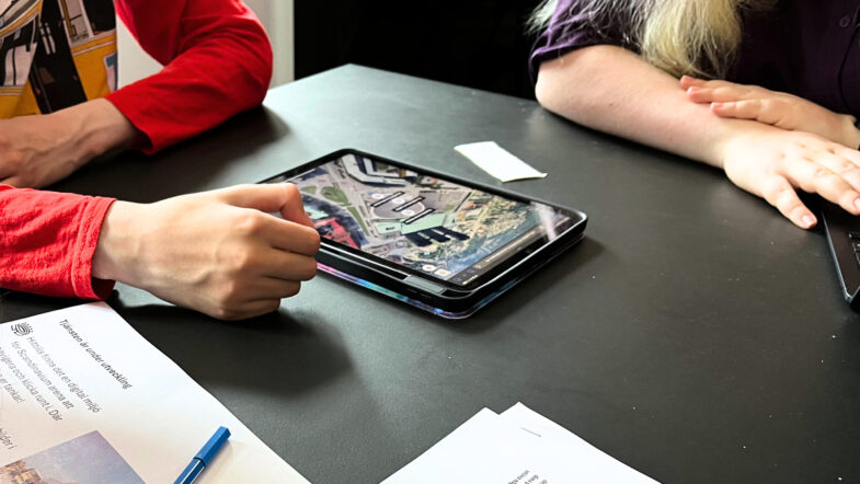 Två personer sitter vid ett bord med en surfplatta som visar en stadskarta. Framför dem ligger papper och en penna.